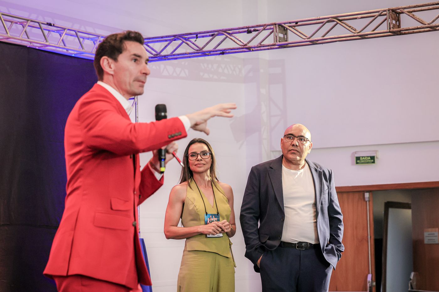 João Adolfo no palco, de terno vermelho, apontando para a plateia